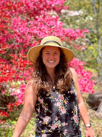 A woman in a wide brimmed hat and floral dress stands in front of blooming trees.